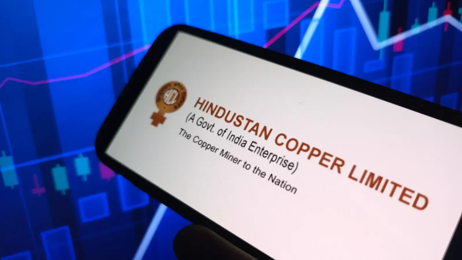 Person holding a copper coin with stock growth graph and Hindustan Copper logo on mobile.