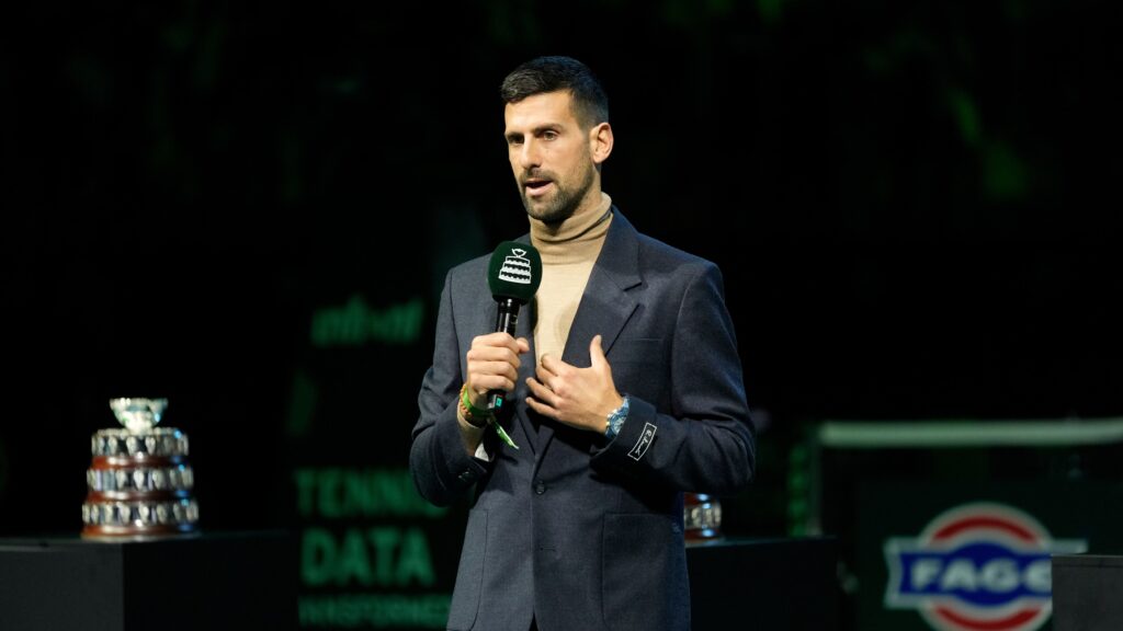 Novak Djokovic speaking at a podium during a tennis event, looking serious.