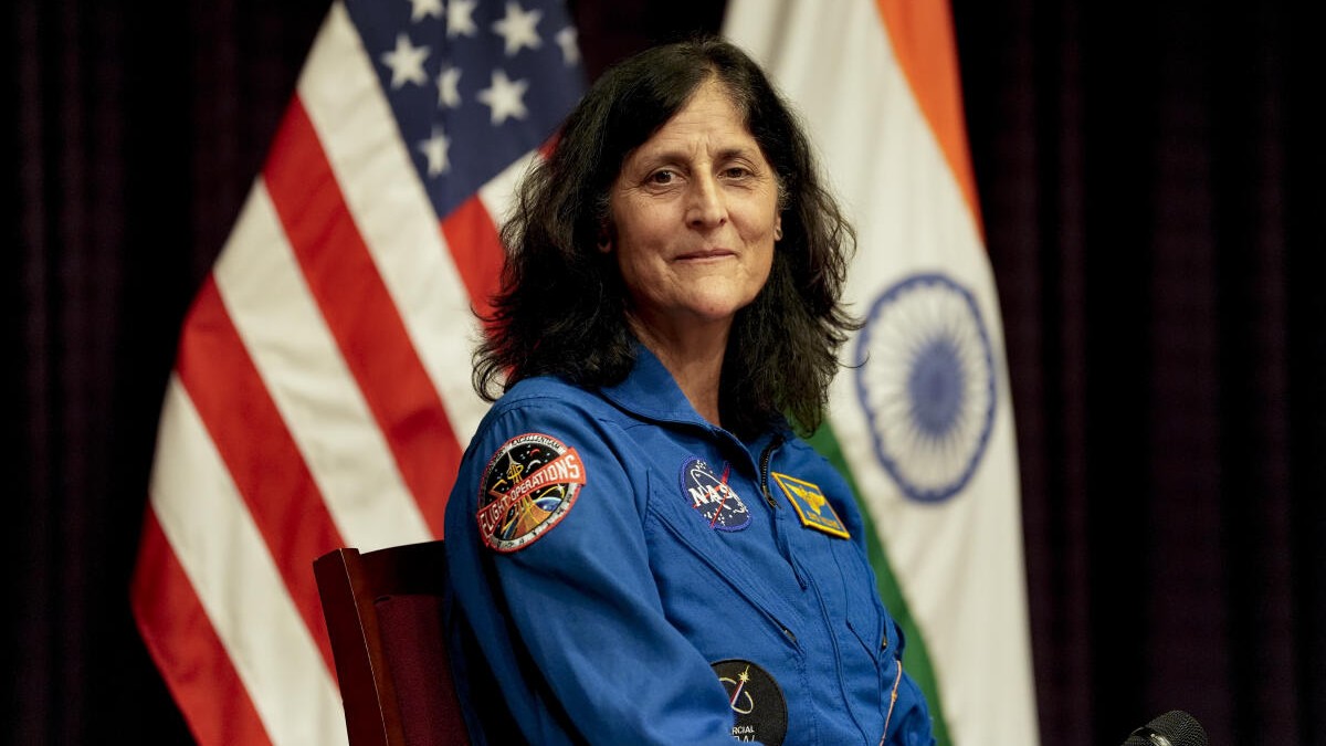 Sunita Williams in blue flight suit sitting in front of US and Indian flags during a press event.
